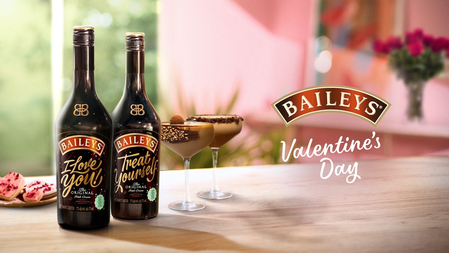 Two Baileys hot chocolates served in a glass, and garnished with chocolate sauce, whipped cream and toasted marshmallow, with a sparkler added for decoration. They sit alongside a bottle of Baileys Original Irish Cream. 